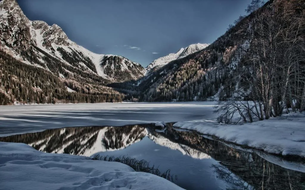 Lago alpino parzialmente ghiacciato con riflessi delle montagne innevate al crepuscolo