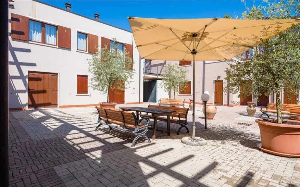 Corte pavimentata con tavolo, panchine in legno e grande ombrellone beige