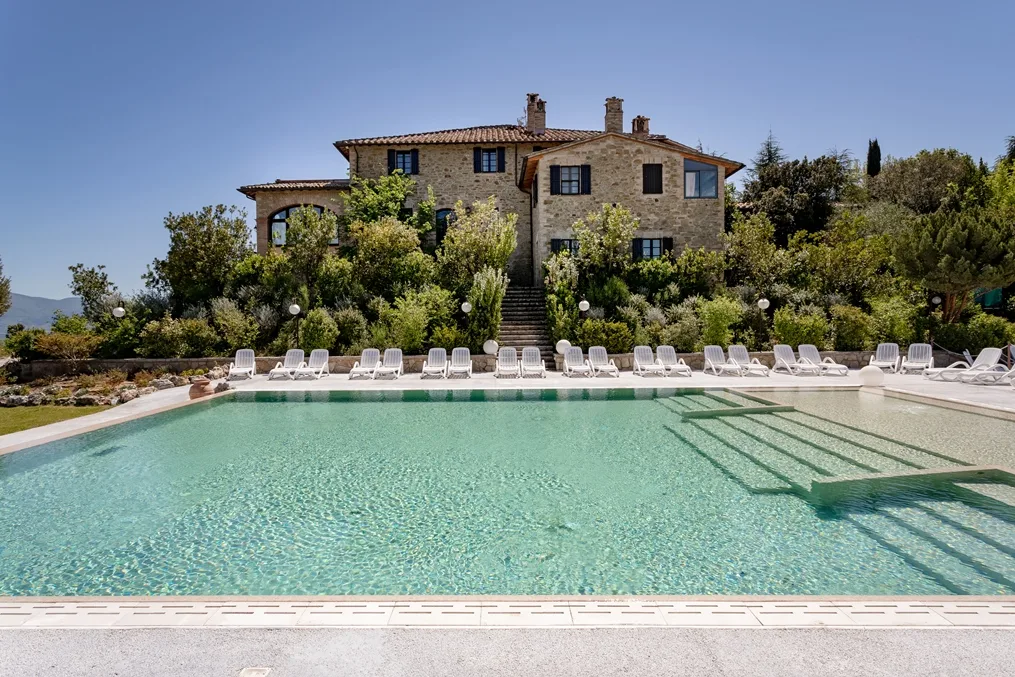 Piscina con acqua cristallina e casale in pietra a due piani sullo sfondo