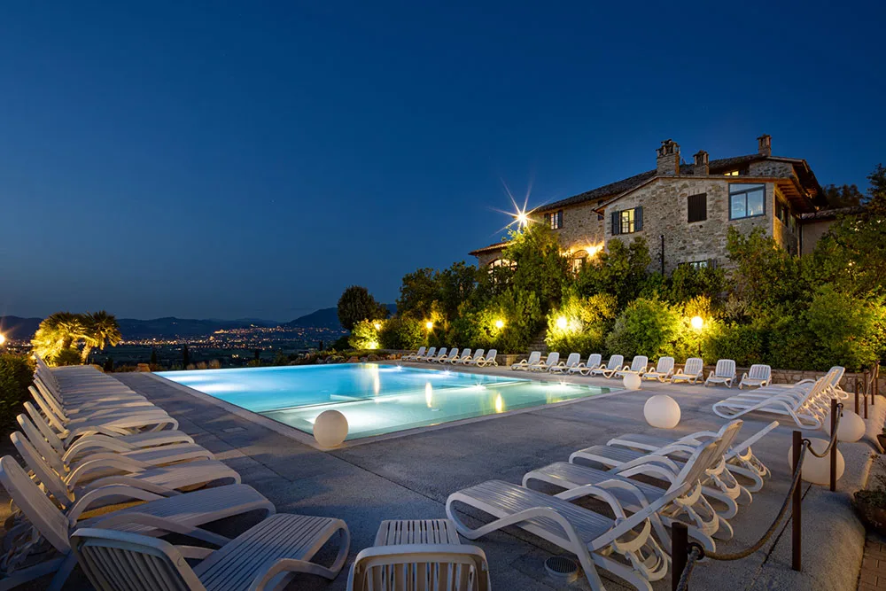Piscina illuminata di sera con lettini bianchi e casale in pietra sullo sfondo