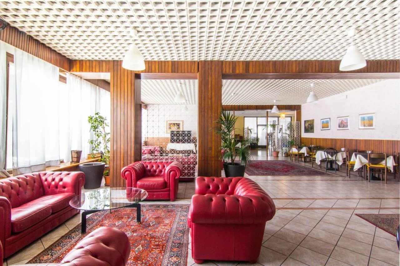 Ampia hall con divani chesterfield in pelle rossa, colonne in legno, piante e sala ristorante sullo sfondo