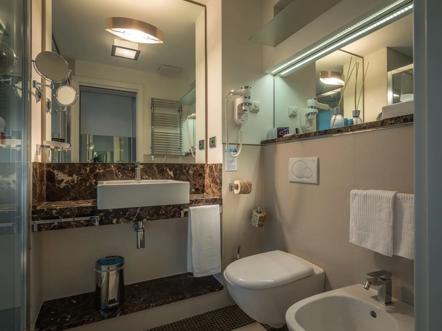 Bagno moderno con lavabo bianco, rivestimenti in marmo, WC, bidet e specchio illuminato