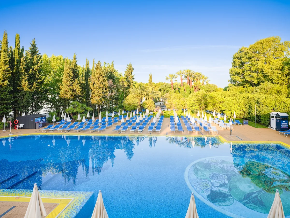 Grande piscina esterna con lettini blu, ombrelloni chiusi e vegetazione rigogliosa