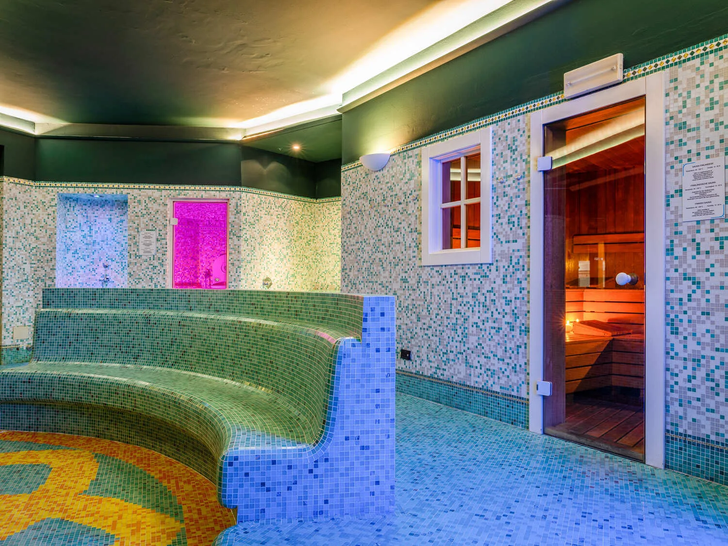Area wellness con mosaico colorato, panche, bagno turco e accesso alla sauna in legno