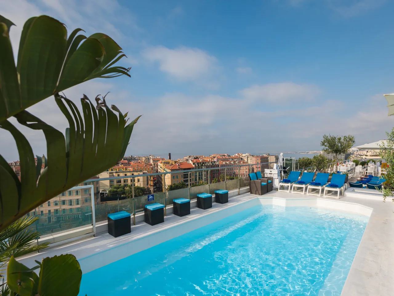 Piscina panoramica sul tetto con lettini blu e vista sui tetti della città e sul mare