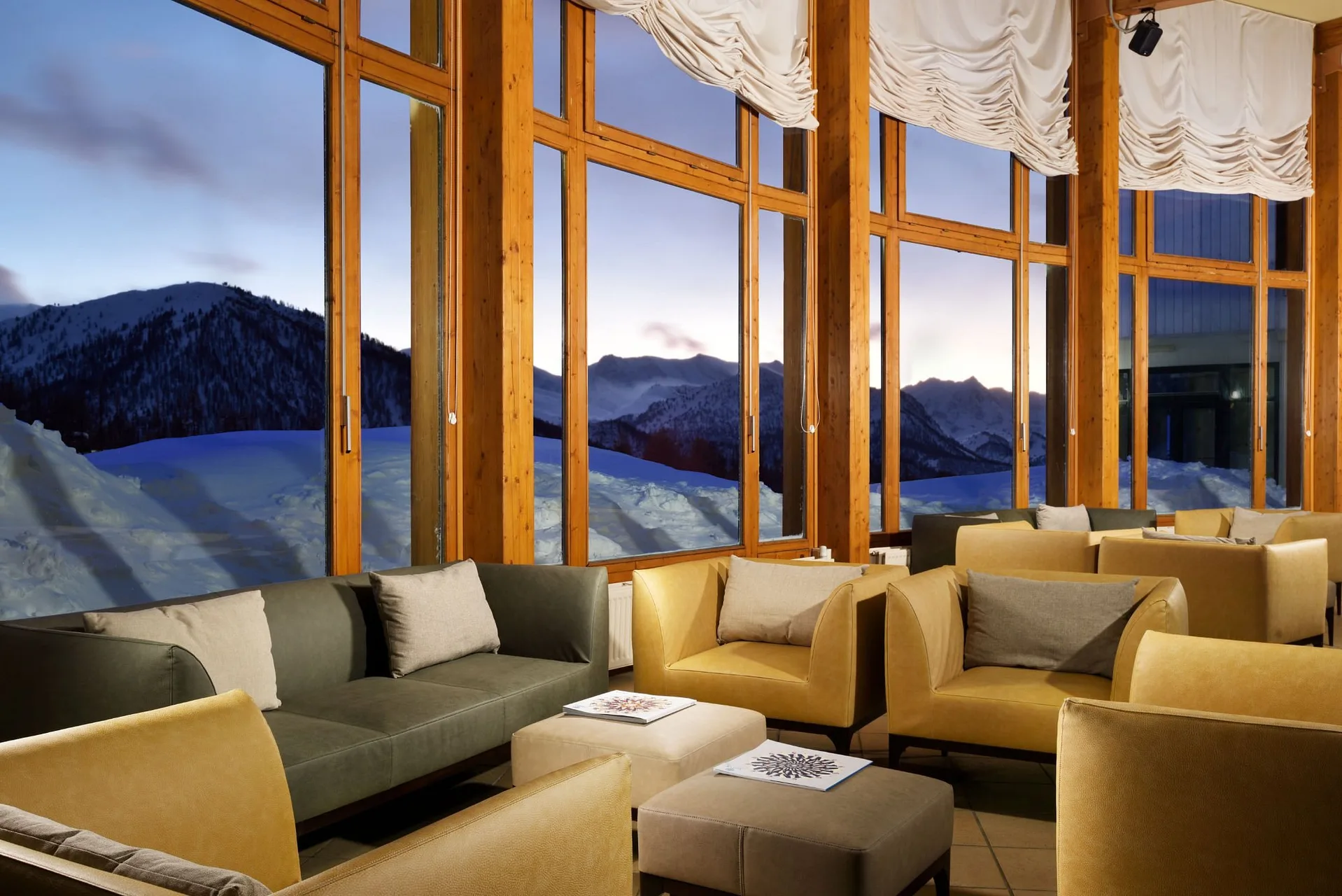 Area lounge con poltrone e divani davanti a grandi vetrate con vista sulle montagne al crepuscolo