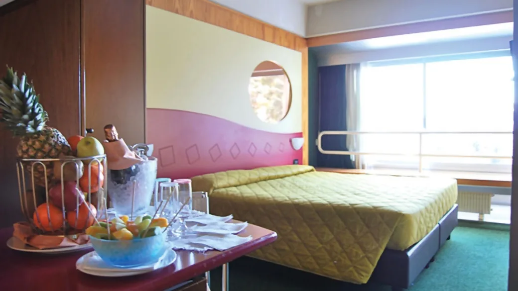 Camera matrimoniale con letto, copriletto verde e tavolo con cesto di frutta e spumante