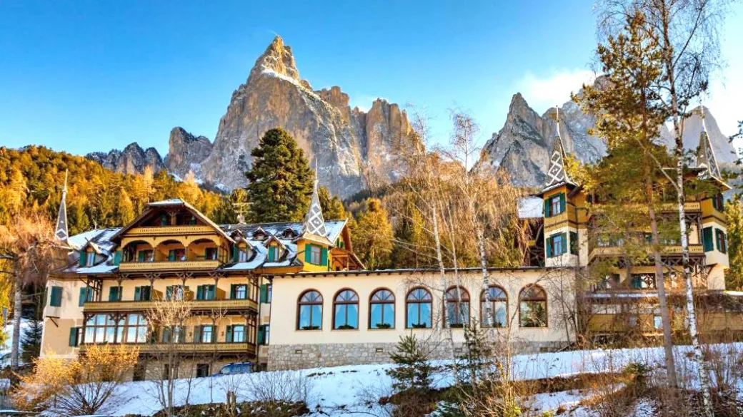 Vista panoramica dell'hotel in stile tirolese con le guglie delle Dolomiti e i boschi autunnali