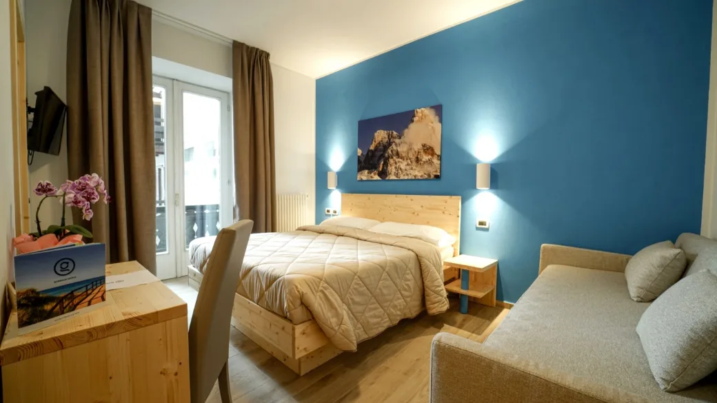 Camera con parete blu, letto matrimoniale e divano, arredata in legno chiaro