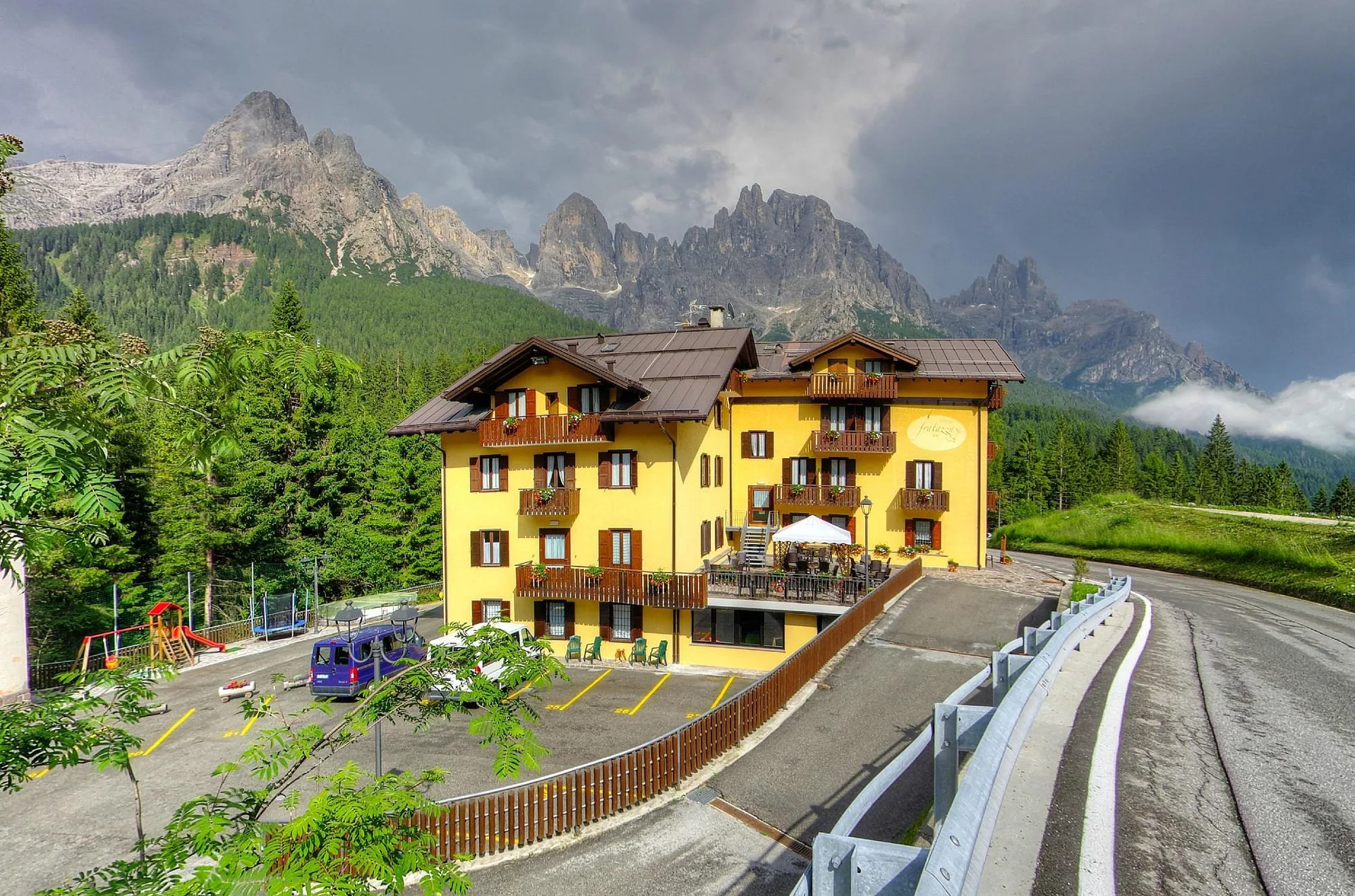 Hotel giallo con balconi fioriti, parco giochi e vista sulle Dolomiti in estate