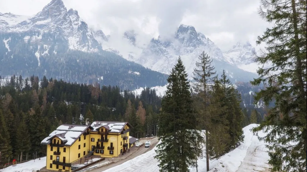 Veduta panoramica dell'hotel giallo tra le conifere con vette dolomitiche avvolte dalle nuvole