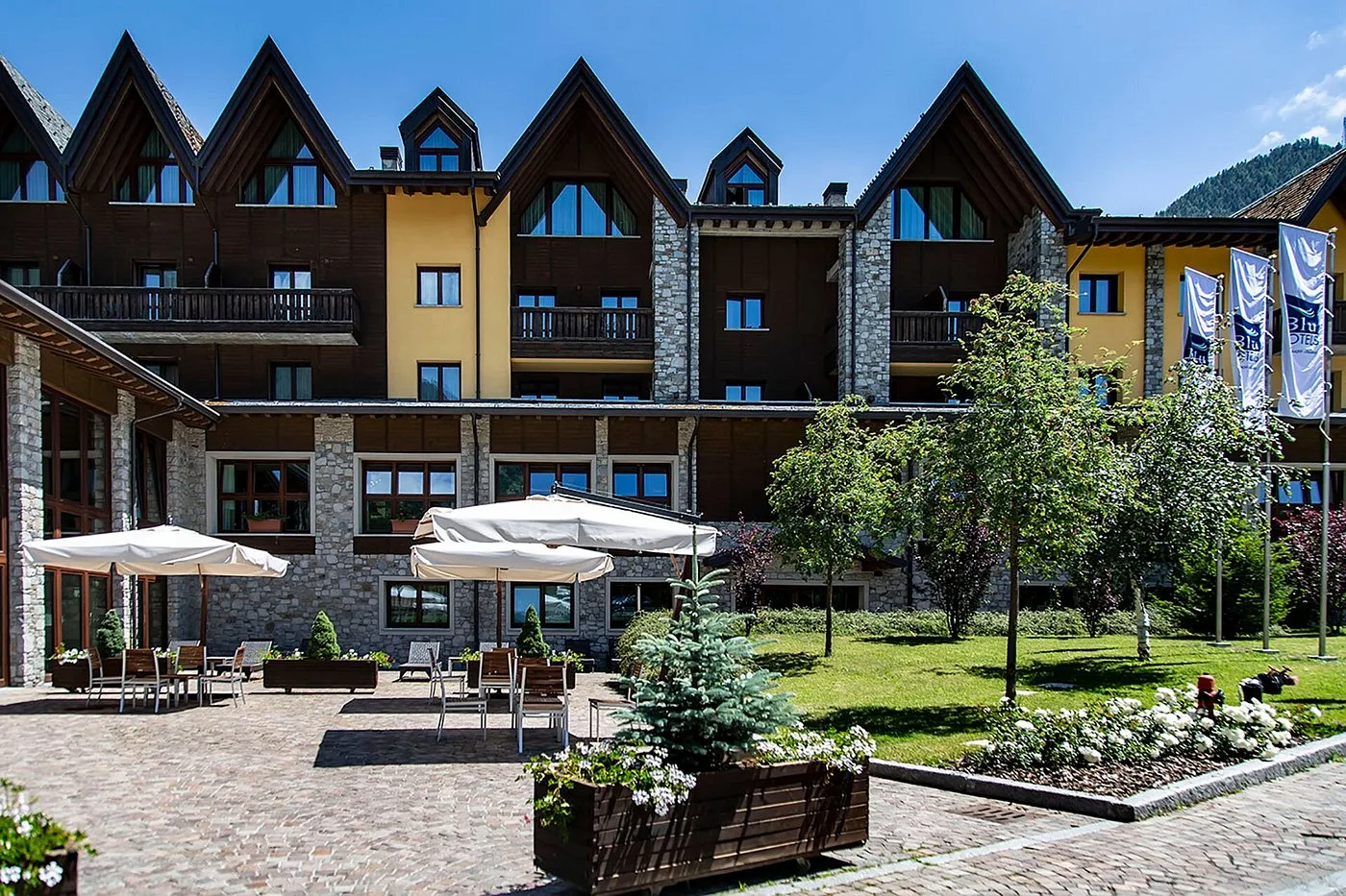 Cortile estivo dell'hotel con ombrelloni, tavoli e giardino curato in stile alpino