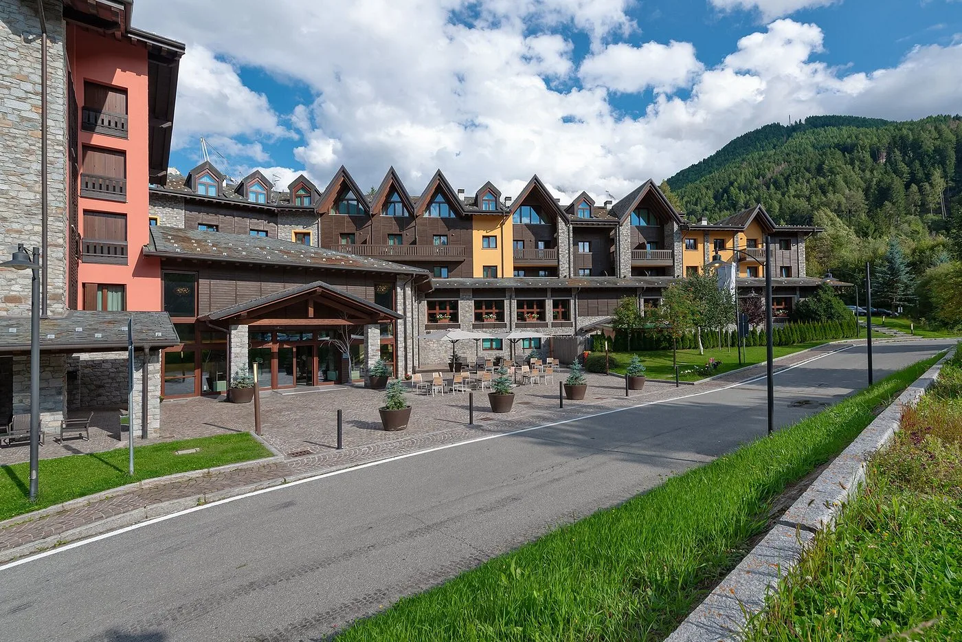 Esterno estivo dell'hotel in stile chalet con terrazza, prati verdi e montagne boscose
