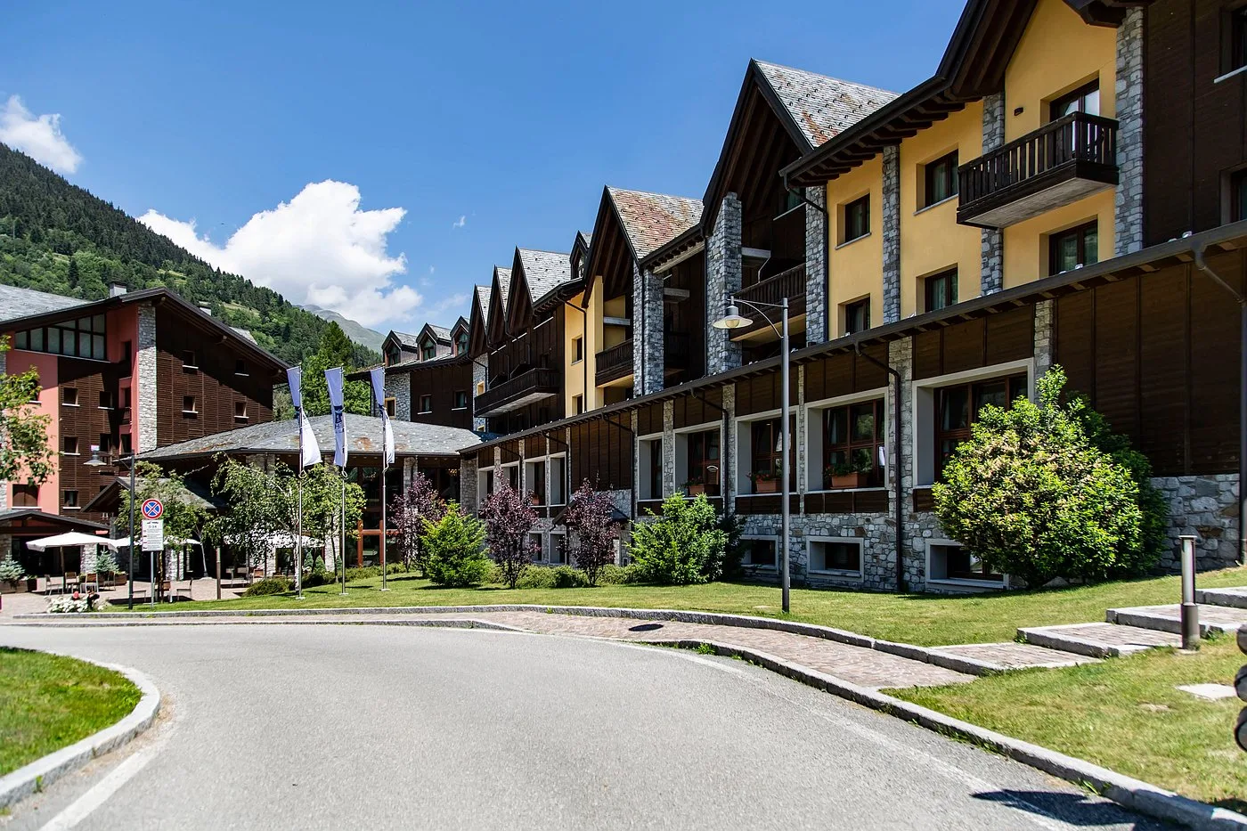 Vista estiva della facciata dell'hotel in stile montano con giardini e strada antistante