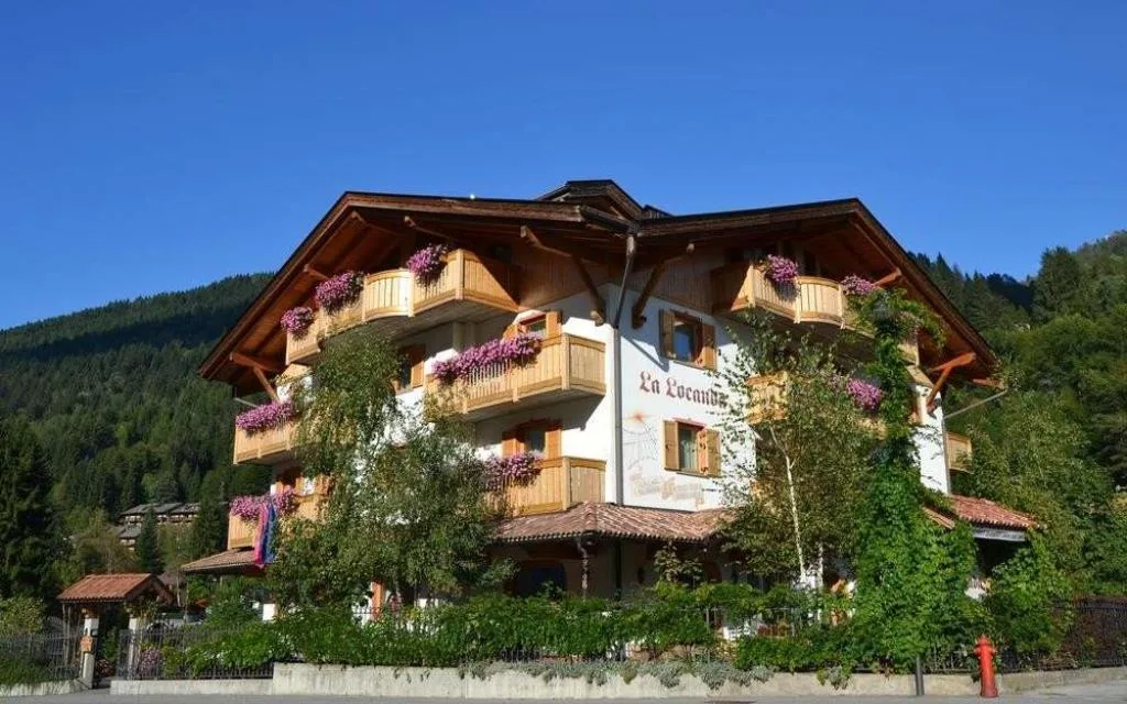 Facciata dell'hotel La Locanda con balconi fioriti e boschi sullo sfondo in estate