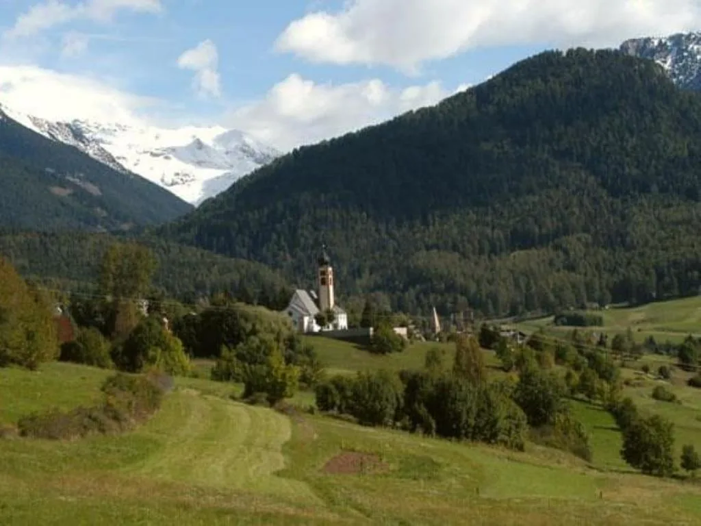 Paesaggio alpino con chiesa, prati verdi e montagne innevate sullo sfondo