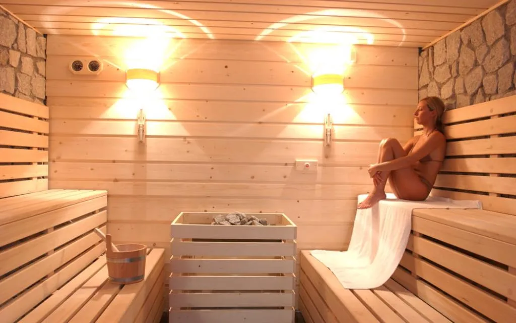 Interno della sauna in legno chiaro con illuminazione calda e persona seduta a rilassarsi