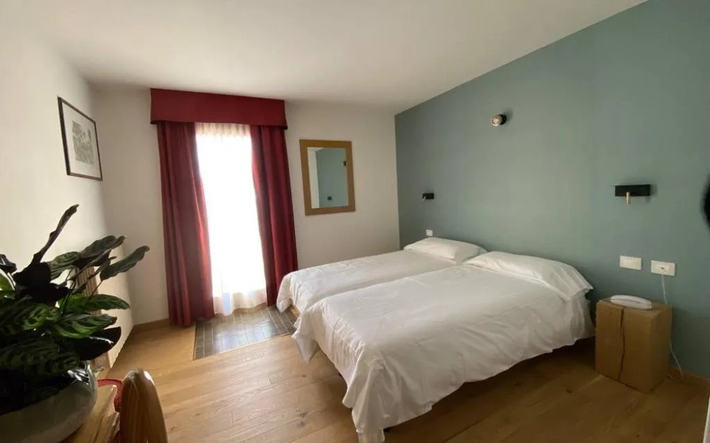Camera doppia con parete verde acqua, letto bianco, tende rosse e pianta decorativa