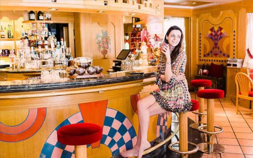Bar dell'hotel con bancone in legno decorato, sgabelli rossi e donna seduta con drink