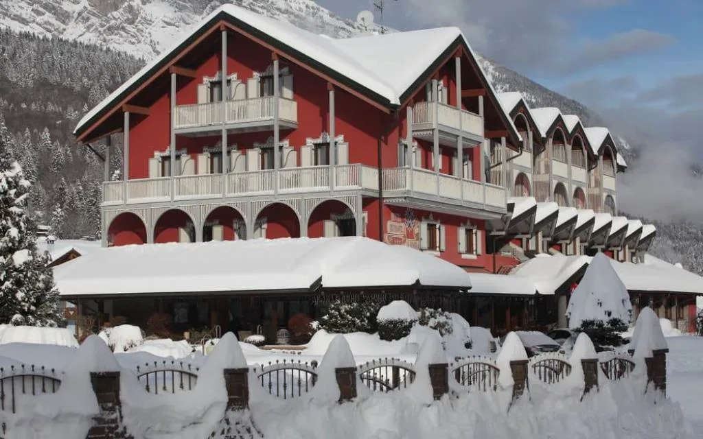 Hotel dalla facciata rossa completamente innevato con montagne e bosco sullo sfondo